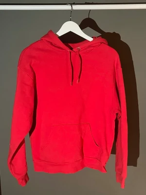 Röd hoodie med magficka - En enkel och stilren röd hoodie med huva och dragsnören. Hoodien har en stor magficka framtill och ribbade muddar vid ärmslut och nederkant. Perfekt för dig som gillar en klassisk look. Materialet känns mjukt och bekvämt, troligen bomullsblandning.