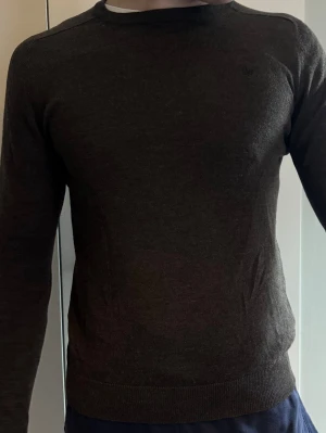 Mörkbrun stickad tröja från Morris  - Mörkbrun stickad tröja med rund hals och diskret broderad  på bröstet. Tröjan har lång ärm och ribbade muddar vid ärmslut och nederkant. Perfekt för lager på lager och enkel att matcha till olika outfits.