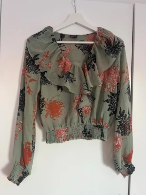 Grön blommig blus med volang - Trendig blus i ljusgrönt med stora blommönster i svart och korallrött. Blusen har volangdetalj framtill, resår i midjan och vid ärmslut samt långa ärmar. Tillverkad i ett lätt, vävt material som ger en luftig känsla. Perfekt för dig som gillar en romantisk och bohemisk stil.