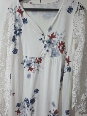 Vit blommig långklänning med spetsärmar - Romantisk vit långklänning med blommigt mönster i blått och rött. Klänningen har v-ringning, dekorativa knappar framtill och långa spetsärmar med vackert mönster. Perfekt för sommarens alla tillfällen.