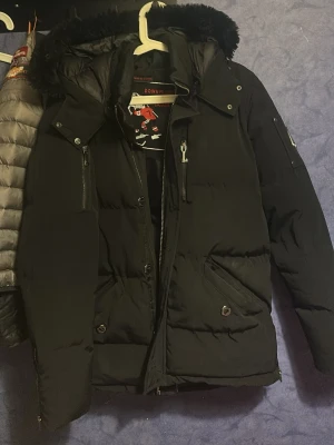 Svart dunjacka från Moose Knuckles - Säljer en svart dunjacka från Moose Knuckles med avtagbar huva och pälskant. Jackan har flera fickor med dragkedja och tryckknappar samt den ikoniska metall-loggan på ärmen. Perfekt för kalla vinterdagar och riktigt snygg streetstil.