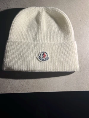 Vit ribbstickad mössa från Moncler( pris kan diskuteras) - Säljer en stilren vit ribbstickad mössa från Moncler med uppvikt kant och klassisk Moncler-logga framtill. Mössan är gjord i mjukt material och har en enkel, clean look som passar till det mesta.