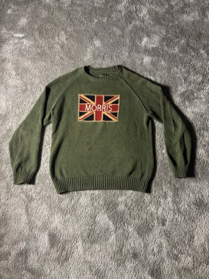 Olivgrön stickad tröja Morris XL - Olivgrön stickad tröja från Morris med Union Jack-flagga och broderad logga på bröstet. Tröjan har rund hals, ribbade muddar och är perfekt för dig som gillar klassisk stil med en twist.