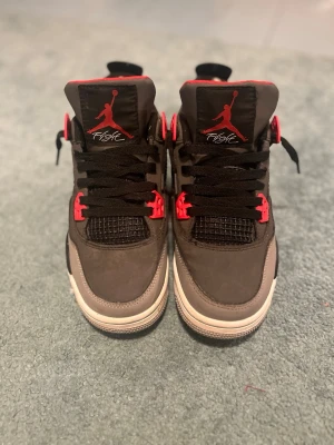 Air Jordan 4 Retro svart/röd - Säljer ett par Air Jordan 4 Retro sneakers i svart och grått med röda detaljer. Skorna har klassisk mesh på sidan, svarta skosnören och vit sula. Ikoniska Jumpman-loggan syns på plösen. Perfekta för dig som gillar streetwear och snygg design.