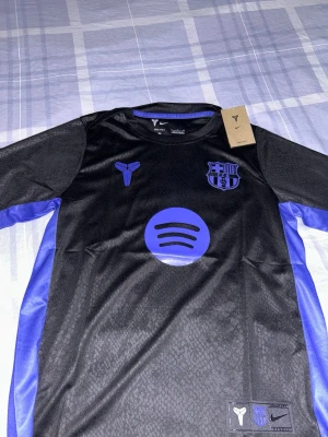 Svart Barcelona tröja Nike Dri-Fit - Snygg svart Barcelona tröja från Nike med lila detaljer och Dri-Fit material. Tröjan har klubbmärke, Kobe Bryant-logga och Spotify-logga på bröstet. Perfekt för fotboll eller träning, med cool design och bekväm passform.