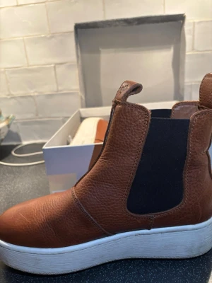 Bruna Chelsea boots från Canada Snow - Säljer ett par snygga Chelsea boots från Canada Snow i brunt läder med elastiska svarta paneler på sidorna och vit platåsula. Skorna har dragflikar både fram och bak för enkel påtagning. Perfekta för dig som vill ha en stilren och modern look.