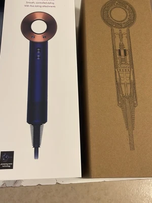 Oanvänd Dyson Supersonic hårfön med tillbehör - Säljer en Dyson Supersonic hårfön i mörkblå och kopparfärg. Medföljer fem stylingtillbehör: styling concentrator, smoothing nozzle, diffuser, gentle air attachment och wide-tooth comb. Modern design i metall och plast, perfekt för olika hårtyper och styling.