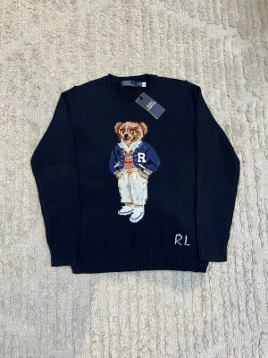 Mörkblå stickad tröja med björnmotiv - Säljer en mörkblå stickad tröja från Ralph Lauren med ett ikoniskt björnmotiv på bröstet. Tröjan har rund hals, ribbade muddar och RL-broderi nedtill. Perfekt för dig som gillar preppy stil och vill ha något unikt i garderoben.