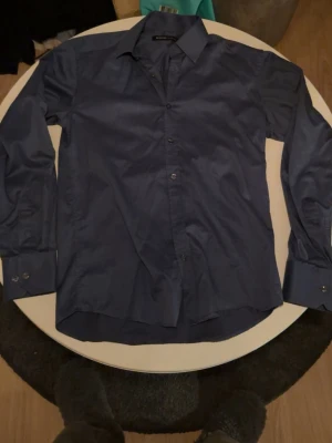 Blå skjorta från Selected Homme - Stilren blå skjorta från Selected Homme med klassisk krage och knappar framtill. Skjortan har lång ärm med manschett och är tillverkad i ett mjukt bomullstyg. Perfekt för dig som gillar en enkel men snygg look.