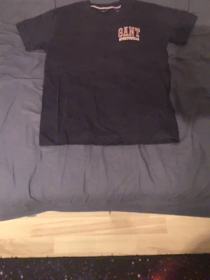 Svart GANT t-shirt med tryck - Säljer en svart t-shirt från GANT med trycket 'GANT SPORTSWEAR' i rött och vitt på bröstet. Klassisk passform och rund halsringning. Perfekt till jeans eller shorts för en enkel och stilren look.