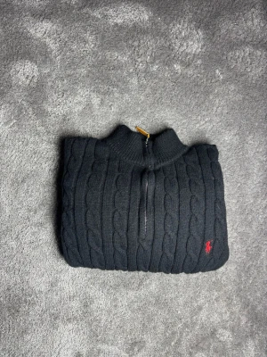 Mörkblå kabelstickad Ralph Lauren M - Säljer en mörkblå kabelstickad tröja från Polo Ralph Lauren med halv dragkedja och röd broderad logga på bröstet. Tröjan har ribbade muddar och hög krage, perfekt för lager på lager. Klassisk och stilren design som funkar till jeans eller chinos.