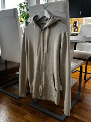 Beige stickad hoodie med dragkedja - Säljer en beige stickad hoodie från zalando med dragkedja framtill och två fickor. Hoodien har ribbade muddar vid ärmslut och nederkant samt justerbar huva med snören. Perfekt för en avslappnad och stilren look. Hör av er vid frågor!