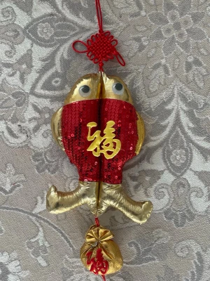 Kinesisk fiskdekoration i guld och rött - Unik hängande dekoration formad som en fisk i guldigt tyg med röda paljetter och kinesiska tecken i gult. Dekorationen har stora ögon, röd snodd för upphängning och en liten guldfärgad påse med rött tecken längst ner. Perfekt för att skapa festlig stämning.
