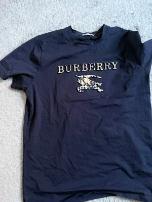 Mörkblå Burberry t-shirt med logga - Snygg mörkblå t-shirt från Burberry med klassisk logga och riddartryck på bröstet. T-shirten har rund hals och korta ärmar, tillverkad i mjuk bomull för skön känsla. Perfekt för dig som vill ha en stilren och exklusiv look.