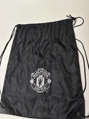 Manchester United svart gympåse - Svart gympåse med Manchester United-logga framtill. Tillverkad i lätt och slitstark syntet med dragskostängning och snören som axelband. Perfekt för träning eller skola för dig som vill visa vilket lag du hejar på.