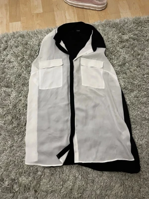 Vit och svart ärmlös blus med fickor - Snygg ärmlös blus i vitt med svarta detaljer och två bröstfickor. Blusen har en klassisk krage och knäppning framtill. Perfekt för en trendig och stilren look.