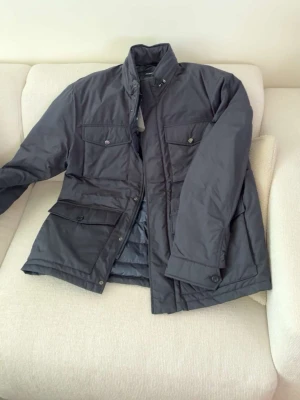 Mörkblå dunjacka från J.Lindeberg - Snygg mörkblå dunjacka från J.Lindeberg med Primaloft-isolering. Jackan har hög krage, fyra framfickor med lock och knapp, samt dragkedja och knappstängning. Perfekt för blåsiga dagar och stilren look. Helt ny med lappen  kvar, hör av er vid några frågor. Lite varmare och passar som sen vinter/ vårjacka NYPRIS: 4200