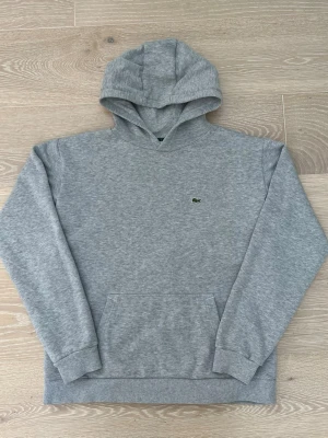 Lacoste Hoodie - Grå - Storlek 176 cm/S - En hoodie från märket Lacoste, i färgen grå med det gröna Lacoste märket. Storlek 176 cm, vilket motsvarar cirka S i vuxen storlek. Skicket är utmärkt, då hoodien är väldigt sparsamt använd. Nypris: 1149kr. Frakt sker inom 24h! Hör gärna av er vid funderingar!🤗
