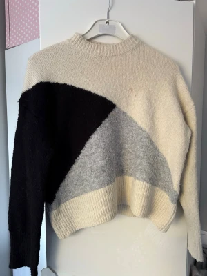 Stickad tröja i beige, svart och grå - Säljer en stickad tröja med cool färgblock-design i beige, svart och ljusgrå. Tröjan har rund halsringning, ribbade muddar och är lite boxig i modellen. Perfekt för dig som gillar en avslappnad och trendig stil.