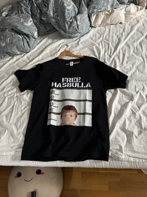 Svart t-shirt med Free Hasbulla-tryck - Cool svart t-shirt med stort tryck på framsidan där det står 'FREE HASBULLA' och en mugshot-inspirerad design. T-shirten har rund hals och korta ärmar, perfekt för en avslappnad streetwear-look.