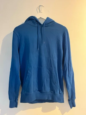 Blå hoodie med magficka - En enkel och stilren blå hoodie med huva och dragsnören. Hoodien har en stor magficka framtill och ribbade muddar vid ärmslut och nederkant. Storlek: S. Aldrig använd. 
