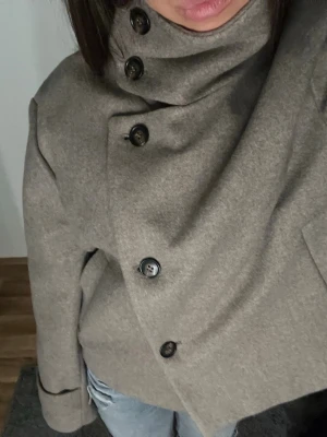Beige ullkappa med hög krage - Snygg beige kappa i ullmix med hög krage och stora svarta knappar i omlottmodell. Kappan har raka ärmar och två sidofickor. Perfekt för dig som vill ha en stilren och modern look till hösten eller vintern.