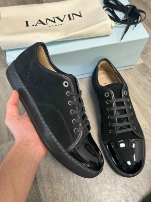 Svarta sneakers från Lanvin - Snygga svarta sneakers från Lanvin med ovandel i mocka och glansig tå i lack. Skorna har svarta skosnören, metallfärgade öljetter och en klassisk låg silhuett. Perfekta för dig som gillar stilrena detaljer och exklusiv känsla. Äkta såklart och helt nya allt på bilderna tillkommer.