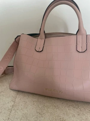 Ljusrosa handväska från Valentino - Snygg ljusrosa handväska från Valentino i croco-mönstrat konstläder. Väskan har två handtag, avtagbar axelrem och guldfärgad logga framtill. Perfekt för dig som gillar stilrena detaljer och trendig design.