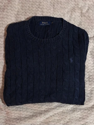 Ralph Lauren - Kabelstickad tröja i mörkblått från Polo Ralph Lauren. Klassisk rund halsringning, långa ärmar och diskret broderad logga på bröstet. Tröjan har ribbade muddar vid ärmslut och nederkant. Perfekt för en stilren och tidlös look. Längd 66 cm & bredd 42 cm 