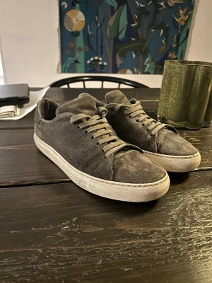 Grå mockasneakers från Axel Arigato - Snygga grå sneakers i mocka från Axel Arigato med vita sulor och klassisk låg profil. Dom är lite smutsiga men det går att få bort. Du får med axel arigsto kartongen också. Dom är köpa från NK och har använts inte jätte mycket men hyfsat mycket i skolan osv. Dom är bara slitna lite grann på sulorna efter man gått på rull grus. Annars är dom i fint skick och inte hål eller något sånt. 