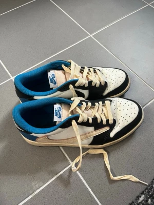 Travis Scott Nike Air Low sneakers svart/vit/blå - Nike Air sneakers i low-modell med svart, vit och beige ovandel, blå insida och vita skosnören. Klassisk swoosh på sidan och perforerad tåbox för extra ventilation. Perfekta för dig som gillar streetwear och sportig stil. Travis Scott