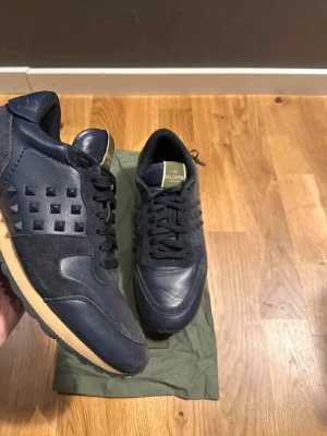 Valentino svarta sneakers med nitar inga defekter. - Snygga svarta sneakers från Valentino med coola nitar på sidorna. Skorna har en mix av läder och textil, svarta skosnören och beige sula. Märkeslogga på plösen i guld. Perfekta för dig som vill sticka ut med en exklusiv touch.