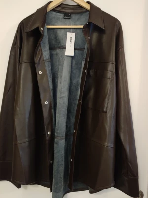 Mörkbrun skinnjacka från Gina Tricot - Stilren mörkbrun skinnjacka från Gina Tricot i oversized modell. Jackan har klassisk krage, tryckknappar framtill och en bröstficka. Utsidan är i slätt konstläder och insidan har mjukt foder. Perfekt för en trendig look.