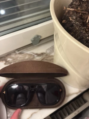 Svarta Tom Ford solglasögon - Säljer ett par svarta solglasögon från Tom Ford med klassisk rektangulär form. Glasögonen har mörka linser och diskreta gulddetaljer vid bågarna. Kommer i ett brunt fodral med Tom Ford-logga i guld. Perfekt accessoar för soliga dagar. Helt äkta 