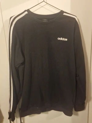 Svart Adidas sweatshirt med vita ränder - Svart sweatshirt från Adidas med klassiska vita ränder längs ärmarna och Adidas-logga på bröstet. Tröjan har rund halsringning och långa ärmar. Perfekt för träning eller chill. Materialet är mjukt och bekvämt, troligen bomullsmix.
