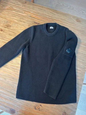 Svart stickad tröja från C.P. Company - Säljer en svart ribbstickad tröja från C.P. Company med deras ikoniska linsdetalj på ärmen. Tröjan har rund halsringning och långa ärmar, perfekt för dig som gillar streetwear och stilrena plagg.