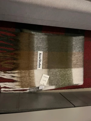 Rutig halsduk från Acne Studios - Säljer en snygg rutig halsduk från Acne Studios i mjuk ullblandning. Färgerna går i jordnära toner som olivgrön, brun, beige, grå och vit. Halsduken har fransar i ändarna och Acne Studios-logga påsydd. Perfekt accessoar för att lyfta din outfit. Orginal pris 3100