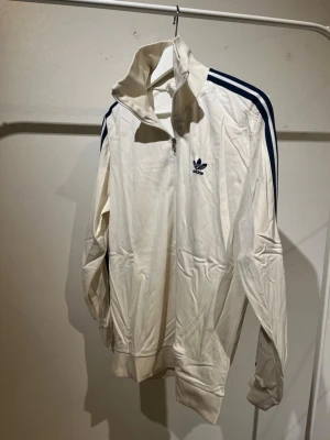 Vit Adidas ziptröja med blå ränder - Säljer en vit ziptröja från Adidas med klassiska blå ränder längs ärmarna och Adidas-logga på bröstet. Tröjan har hög krage, dragkedja framtill och ribbade muddar. Perfekt för en sportig och avslappnad stil.