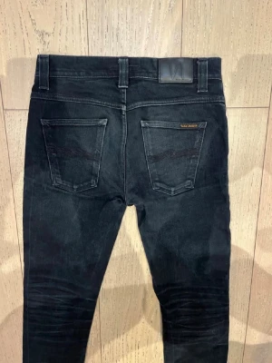Nudie grim tim - Säljer ett par svarta slim fit jeans från Nudie Jeans med klassisk femficksmodell och diskreta slitningar. Jeansen har svarta sömmar, läderpatch bak i midjan och är tillverkade i mjuk bomullsdenim med stretch. Perfekta för en clean och stilren look. W30 L32