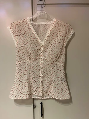 Vit blommig blus med knappar - Söt vit blus med små röda blommor, v-ringning och dekorativa knappar framtill. Blusen har spetsdetaljer längs kanterna och är tillverkad i ett tunt material. Aldrig använd 