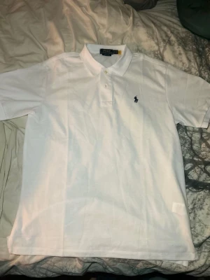 Vit pikétröja från Polo Ralph Lauren - Klassisk vit pikétröja från Polo Ralph Lauren med korta ärmar och den ikoniska broderade loggan på bröstet. Tröjan har krage och knappar framtill, perfekt för en clean och stilren look.