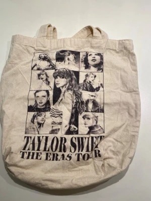 Taylor Swift The Eras Tour tygpåse - Beige tygpåse med tryck från Taylor Swift The Eras Tour. Ena sidan har collage med svartvita bilder och text, andra sidan har texten 'Taylor Swift The Eras Tour VIP'. Perfekt för fans och som accessoar till vardagen. Påsen är tvättad och blivit mindre än originalstorleken 