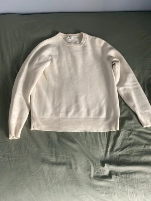 Uniqlo - Säljer en stilren kritvit stickad tröja med rund hals och långa ärmar. Tröjan har en enkel, clean design utan mönster och är ribbstickad vid ärmslut och nederkant. Perfekt för dig som gillar minimalistisk stil.
