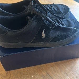 Svarta sneakers från Polo Ralph Lauren - Säljer ett par svarta sneakers från Polo Ralph Lauren med klassisk logga på sidan. Skorna har snörning, rund tå och platt sula. Perfekta för en stilren och avslappnad look. Levereras i originalkartong. Skorna är använda 2-3 gånger lite smutsiga men går att rengöra 