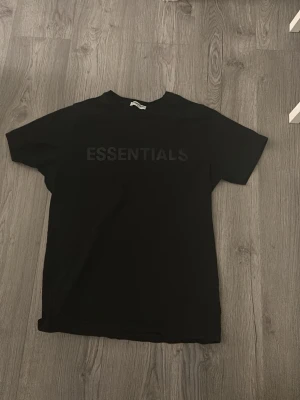Svart Essentials t-shirt - Säljer en svart t-shirt från Essentials med diskret tryck på bröstet. T-shirten har klassisk passform, rund halsringning och är tillverkad i mjukt bomullsmaterial. Perfekt för en clean och stilren look.