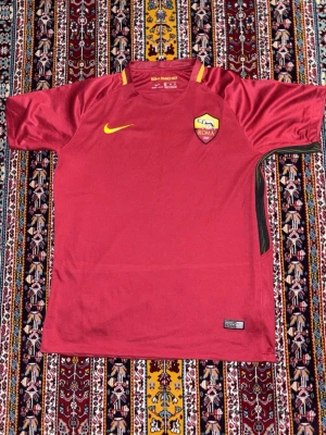 AS Roma matchtröja Nike röd - Säljer en röd AS Roma matchtröja från Nike med klubbmärke på bröstet och gul Nike-logga. Tröjan har korta ärmar, rund hals och gula detaljer vid kragen. Tillverkad i lätt och ventilerande funktionsmaterial, perfekt för fotbollsträning.