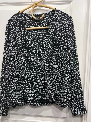 Svartvit mönstrad omlottblus från H&M - Svartvit omlottblus från H&M med abstrakt grafiskt mönster. Blusen har långa ärmar och en avslappnad passform med omlottdesign framtill. Tillverkad i ett mjukt, lätt material som känns skönt mot huden. Perfekt för dig som gillar statementplagg.