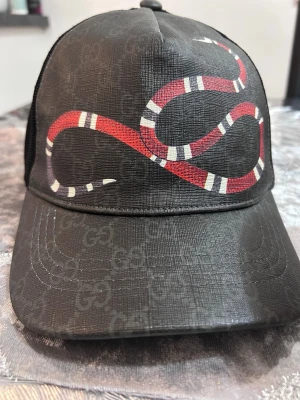 Gucci keps med orm och GG-mönster - Snygg svart keps från Gucci med klassiskt GG-mönster över hela kepsen och en stor röd orm broderad framtill. Kepsen har böjd skärm och mesh på sidorna för extra ventilation. Perfekt för dig som vill sticka ut med en unik accessoar.