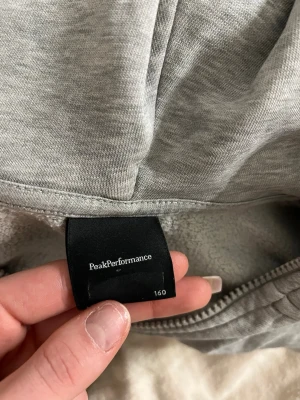 Grå hoodie från Peak Performance - Säljer en ljusgrå hoodie från Peak Performance med dragkedja framtill och två fickor. Hoodien har ribbade muddar vid ärmslut och nederkant samt en broderad logga på bröstet. Tillverkad i mjukt bomullsmaterial och har en klassisk huva.