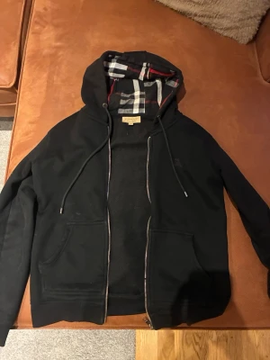 Svart hoodie från Burberry(dragkedjan är sönder) - Snygg svart hoodie från Burberry med dragkedja och klassiskt rutigt foder i huvan. Hoodien har två fickor framtill och justerbara snören i huvan. Perfekt för dig som gillar stilrena plagg med exklusiva detaljer. Dragkedjan är sönder men går och få fixat hos en skräddare var som helst. Det är en anledning till att den säljs billigt.😊